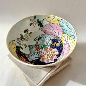 May include: A decorative ceramic bowl with a floral and leaf design in pink, yellow, blue, and green. The bowl is resting on a stack of books with the text "RENOIR JUAULT".