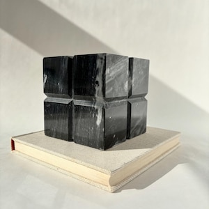 May include: A black and white marble sculpture in a cube shape, resting on a beige book. The sculpture has a geometric design with visible veining. The book has a red spine and a light-colored cover.