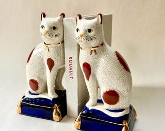 May include: Pair of ceramic cat bookends. The cats are white with brown spots and sit on blue cushions with gold accents. They have gold collars and are positioned on either side of a book with the text "ROUAULT".