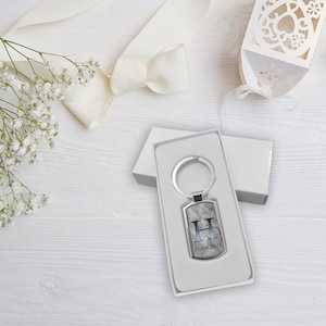 May include: A silver keyring with a rectangular tag featuring the letter "H" and the name "HAROON" engraved on it. The keyring is in a white box.