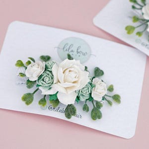 Op de afbeelding: Haarclip met bloemen, met een grote ivoorkleurige roos omringd door kleinere groene en ivoorkleurige rozen en groene bladeren. De clip zit op een witte kaart met de tekst "lila bow" en de Instagram-handle "@lilabowhair".