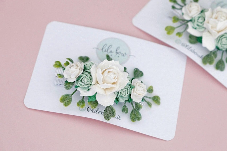 Op de afbeelding: Haarclip met bloemen, met een grote ivoorkleurige roos omringd door kleinere groene en ivoorkleurige rozen en groene bladeren. De clip zit op een witte kaart met de tekst "lila bow" en de Instagram-handle "@lilabowhair".