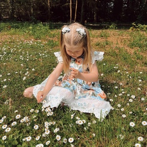 Op de afbeelding: Een jong kind in een bloemenjurk en een witte kanten broek zit in een veld met madeliefjes. De jurk heeft een blauw, oranje en wit bloemenpatroon. Het kind draagt twee witte bloemenhaarclips.