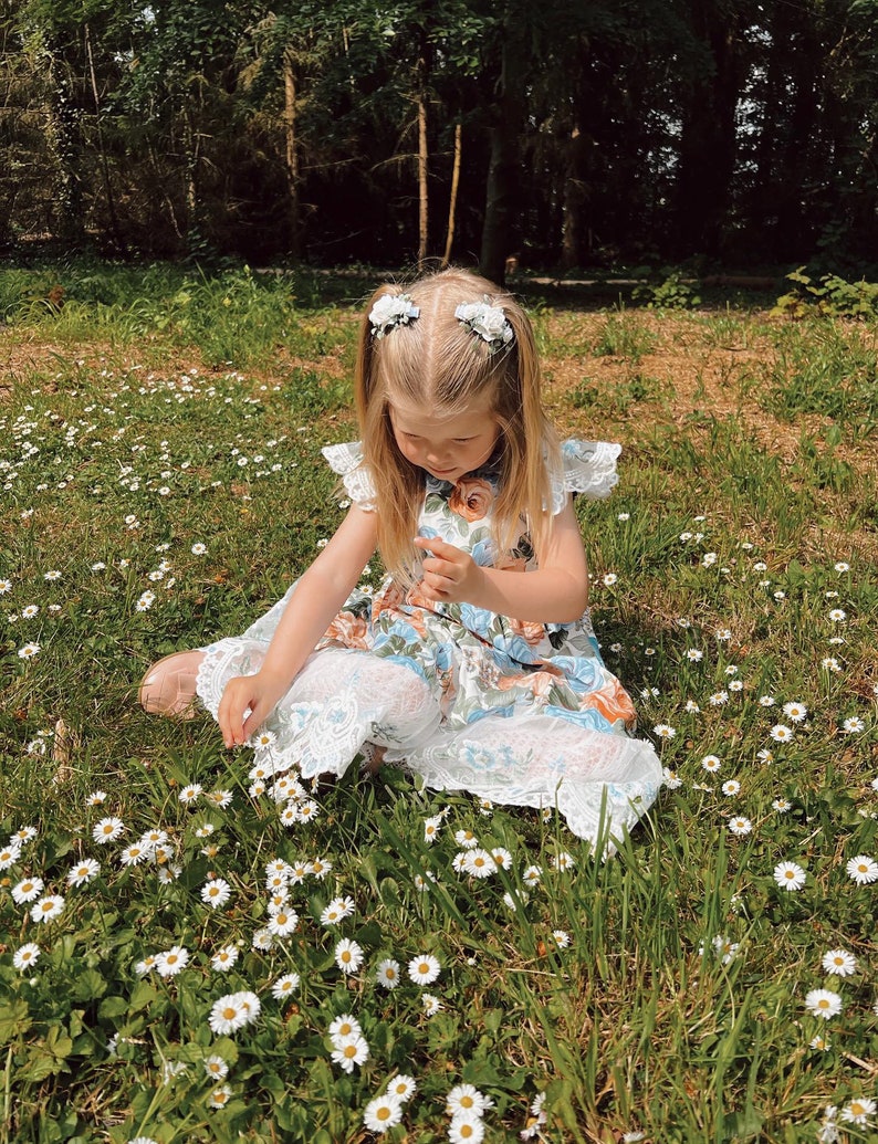 Op de afbeelding: Een jong kind in een bloemenjurk en een witte kanten broek zit in een veld met madeliefjes. De jurk heeft een blauw, oranje en wit bloemenpatroon. Het kind draagt twee witte bloemenhaarclips.