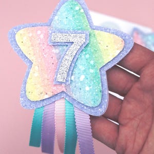 May include: A star-shaped birthday badge with a rainbow glitter design and the number 7 in silver glitter. The badge has a light blue border and is decorated with teal, lavender, and pink ribbons, ideal for a seventh birthday celebration.