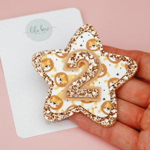 May include: A star-shaped hair clip with a glittery gold border and a lion pattern. The number "2" is in the center. The clip is on a white card with the text "lila boo" in cursive.