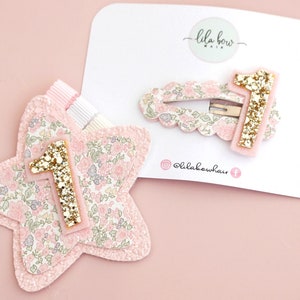 Könnte beinhalten: Eine sternförmige Haarspange und eine gewellte ovale Haarspange, beide mit rosa Blumenmuster und einer goldenen Glitzer-Eins. Die Spangen sind auf einer weißen Karte mit dem Text lila bow hair und dem Social-Media-Handle lilabowhair befestigt.