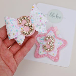 Könnte beinhalten: Eine weiße Glitzer-Haarschleife mit bunten Konfetti-Akzenten und einer goldenen Glitzer-Zahl 3, kombiniert mit einem sternförmigen, rosa-weißen Glitzer-Haaraccessoire mit goldener Glitzer-Zahl 3, befestigt auf einer markierten Präsentationskarte.