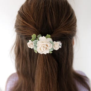Op de afbeelding: Haarclip met cr&egrave;mekleurige rozen en lichtgroene bloemen, met groene bladeren. Het haaraccessoire is geplaatst tegen een achtergrond van bruin haar.