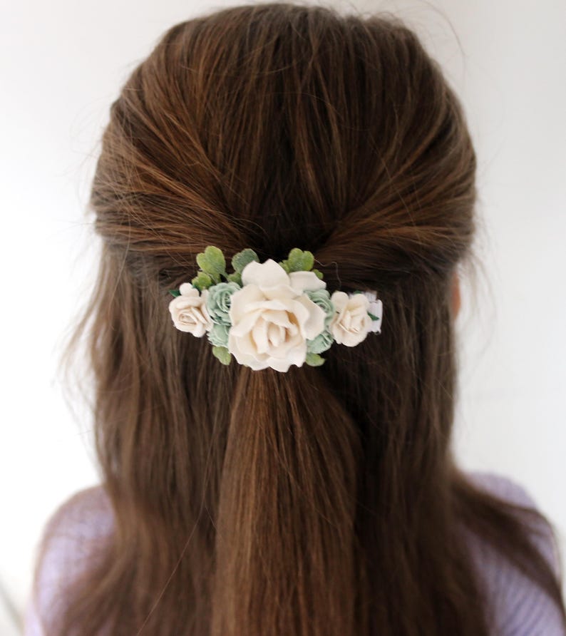 Op de afbeelding: Haarclip met cr&egrave;mekleurige rozen en lichtgroene bloemen, met groene bladeren. Het haaraccessoire is geplaatst tegen een achtergrond van bruin haar.