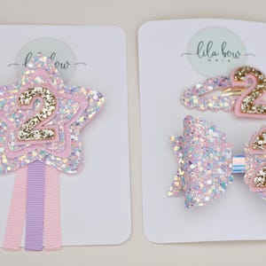 May include: A set of hair accessories on two white display cards labelled Lila Bow Hair. One card features a star-shaped glitter hair clip with a gold glitter number 2 and three ribbon streamers. The other card holds a glitter bow and a snap clip, both featuring a gold glitter number 2.