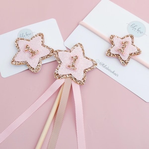 Könnte beinhalten: Rosa sternförmige Haaraccessoires mit goldfarbenem Glitzerrand und der Zahl 1 in der Mitte. Die Accessoires befinden sich auf einem rosa Hintergrund mit einer weißen Karte und einem rosa Haarband. Für einen ersten Geburtstag.