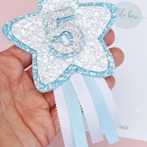 Op de afbeelding: Een stervormige haarclip voor een vijfde verjaardag, met een glitterig lichtblauw en wit ontwerp. Het nummer 5 is in zilveren glitter. Witte en lichtblauwe linten hangen aan de onderkant. Een feestelijk haaraccessoire.