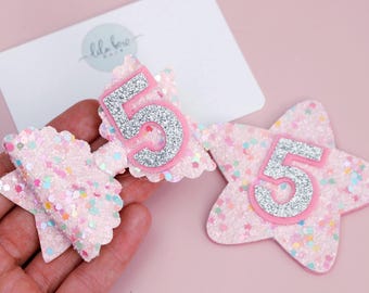 May include: Pink glitter hair bows and star-shaped hair accessory with the number 5 in silver glitter. The bows and star are on a pink background. The brand name "lila bow" is on a white card.