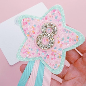 Könnte beinhalten: Haarspange in Sternform mit rosa Glitzerbasis, hellgrünem Rand und einer silbernen Acht in der Mitte. Die Spange hat rosa und blaue Bänder, die von unten herabhängen. Ein festliches Accessoire.