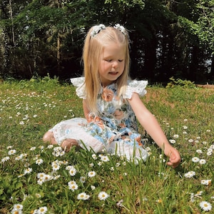 Op de afbeelding: Een jong kind in een bloemenjurk met kanten rand zit in een veld met madeliefjes. De jurk heeft een blauw en oranje bloemenpatroon. Het kind heeft blond haar en draagt ​​haaraccessoires met bloemen.