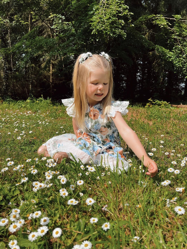 Op de afbeelding: Een jong kind in een bloemenjurk met kanten rand zit in een veld met madeliefjes. De jurk heeft een blauw en oranje bloemenpatroon. Het kind heeft blond haar en draagt ​​haaraccessoires met bloemen.
