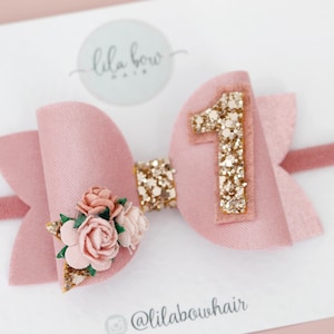May include: A pink bow with a gold glitter number one and a cluster of pink flowers. The bow is attached to a pink elastic headband. The headband is for a baby's first birthday.