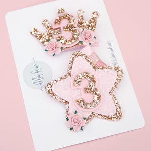 Könnte beinhalten: Zwei glitzernde Haarspangen, eine in Form einer Krone und die andere in Form eines Sterns, beide mit der Zahl "3". Die Krone und der Stern sind rosa mit goldenem Glitzer und kleinen rosa Rosen. Die Spangen befinden sich auf einer weißen Karte.