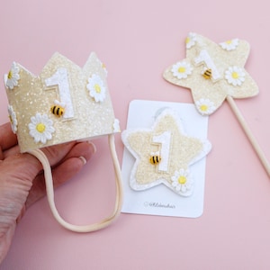 May include: A glittery gold crown with a white number one, white daisy flowers, and small bee accents, paired with a matching star-shaped hair clip and a star-shaped wand on a stick, both featuring the number one, daisies, and bee details.