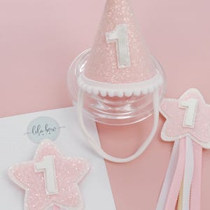 May include: A pink glitter party hat with the number 1, a white pom-pom, and a clear dummy. A star-shaped hair accessory with the number 1 and ribbons. The items are on a pink background. The brand name "lila bow" is visible.