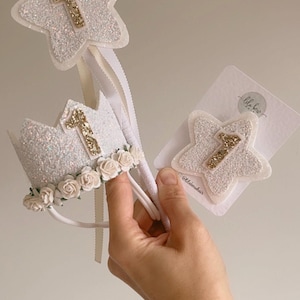 May include: A collection of sparkly white accessories for a first birthday. Includes a star wand, a crown with floral details, and a star hair clip, all with the number 1 in gold glitter.