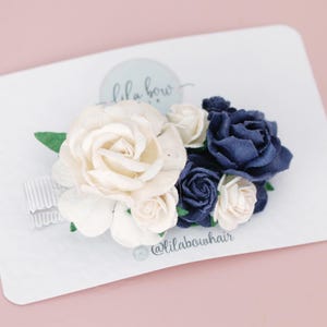 Pode incluir: Um prendedor de cabelo com flores brancas e azul marinho, uma fita branca e o texto "lila bow" e "@lilabowhair".