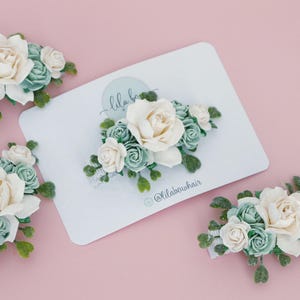 Op de afbeelding: Verschillende bloemen haarclips met witte en zeegroene rozen en groene bladeren zijn gerangschikt op een witte kaart en een roze achtergrond. De kaart heeft de tekst "lila bow" en de Instagram-handle "@lilabowhair".