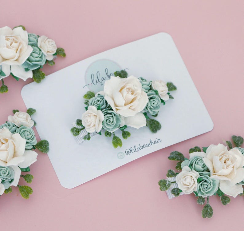 Op de afbeelding: Verschillende bloemen haarclips met witte en zeegroene rozen en groene bladeren zijn gerangschikt op een witte kaart en een roze achtergrond. De kaart heeft de tekst "lila bow" en de Instagram-handle "@lilabowhair".