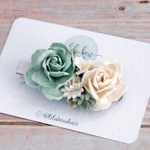 May include: A floral hair clip with a sage green rose and a cream-coloured rose, with small white and green accents. The clip is attached to a light grey striped barrette. The product is displayed on a white card with the text "lila bow" and the Instagram handle "@lilabowhair".