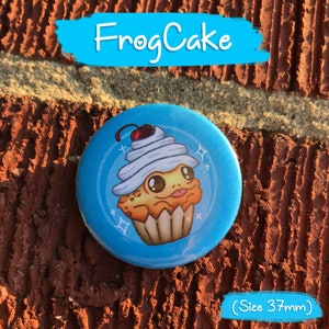 May include: A round, light blue button with the text "FrogCake" in white. The button features a cartoon cupcake with a frog face, white frosting, and a cherry on top. The button is 1.45 inches in diameter.
