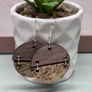 May include: A pair of wooden earrings with a bird and floral design. The earrings are circular, with a dark brown wood top and a printed bottom section. The printed section features a beige background with a bird perched on a branch. Silver-colored ear wires complete the design.