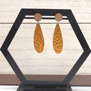 Puede incluir: Pendientes de lágrima de color amarillo mostaza con un patrón de encaje detallado. Los pendientes tienen un perno de madera en la parte superior. Los pendientes se exhiben en un soporte hexagonal negro.
