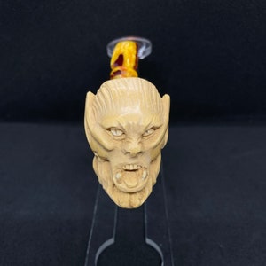Puede incluir: Una pipa de madera tallada a mano con una cara de monstruo detallada y gruñona. La pipa está hecha de madera marrón claro y tiene una boquilla roja y amarilla.