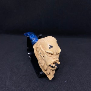 Puede incluir: Una pipa de madera tallada a mano con un diseño de rostro detallado. La pipa está pintada en beige y negro, con una boquilla azul y blanca.