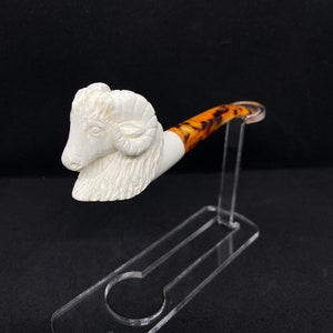 May include: A white carved ram's head pipe with a brown and white stem. The pipe is displayed on a clear acrylic stand.