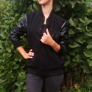 Może przedstawiać: Czarna kurtka typu varsity z czarnymi skórzanymi rękawami i czarnym żebrowanym kołnierzem, mankietami i dołem. Kurtka ma zapinany na guziki przód i dużą literę "M" na piersi. Noszona z czarnym topem i ciemnymi dżinsami.