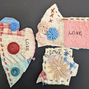 May include: Three handmade fabric hearts with the word "LOVE" stitched on each. The hearts are made of different fabrics and colors, including pink, blue, white, and red. The hearts are decorated with buttons and flowers.