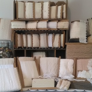 May include: A wooden shelf unit filled with spools of white lace trim. The shelf unit is against a white wall.
