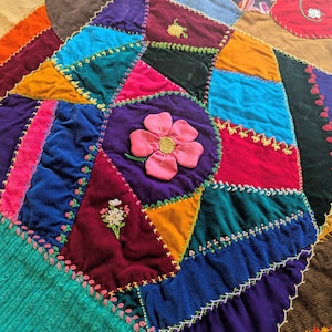 May include: A colorful patchwork quilt with a variety of jewel-toned velvet fabrics. The quilt features embroidered floral details and decorative stitching in white, pink, and yellow thread. The quilt is a mix of geometric shapes and floral designs.