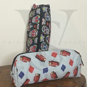 May include: Two pencil cases, one with a black background and a pattern of colorful sugar skulls, and the other with a light blue background and a pattern of red cartoon race cars.