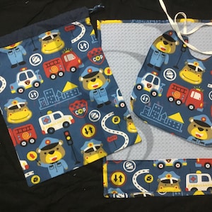 May include: A blue and yellow baby bib, a blue and yellow burp cloth, and a blue and yellow drawstring bag. All three items feature a repeating pattern of cartoon police officers, fire trucks, and ambulances. The text "COP" appears on the items.
