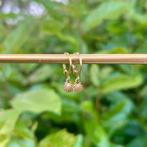 May include: A pair of gold-colored hoop earrings with small seashell charms. The earrings are hanging on a gold bar against a blurred green background of foliage. The earrings are a delicate jewelry item.