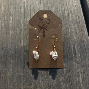 May include: A pair of seashell earrings with gold-colored hooks and small white pearls. The earrings are displayed on a brown tag with the initials "E J" and a palm tree graphic.
