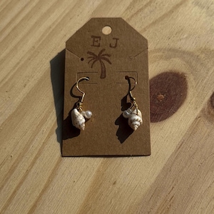 May include: A pair of seashell earrings with gold-colored hooks and small white pearls. The earrings are displayed on a brown tag with the initials "EJ" and a palm tree design. The background is a light brown wooden surface.