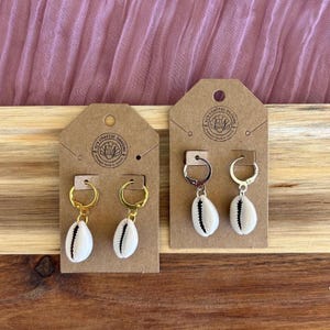 May include: Two pairs of seashell earrings displayed on brown tags. One pair features gold hoops, the other silver. Each earring has a white cowrie shell with a black stripe. The tags have a circular logo with text.