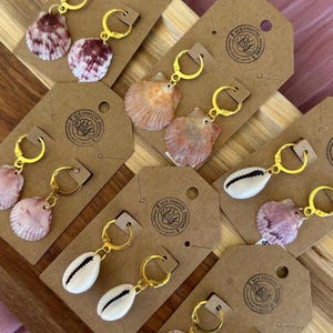 May include: A collection of seashell earrings with gold-colored hoops, displayed on brown tags. The earrings feature various shells in shades of pink, white, and orange. The tags have a circular logo with text. The earrings are designed for a coastal theme.