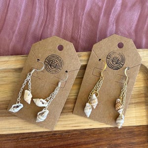 May include: Two pairs of seashell earrings displayed on brown tags. One pair features silver chains and hooks, while the other has gold chains and hooks. Each earring has multiple small seashells dangling from the chains. The tags have a circular logo with text.