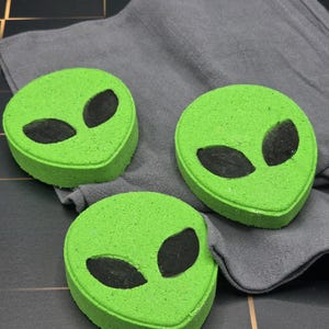 Puede incluir: Tres bombas de baño verdes con forma de alienígena y ojos ovalados negros descansan sobre un paño gris. Las bombas de baño tienen una superficie texturizada y están dispuestas sobre una superficie oscura y embaldosada.