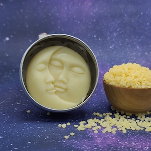 May include: A round tin container with a white, sculpted moon and face design. The container is sitting on a purple background with yellow wax beads scattered around it.